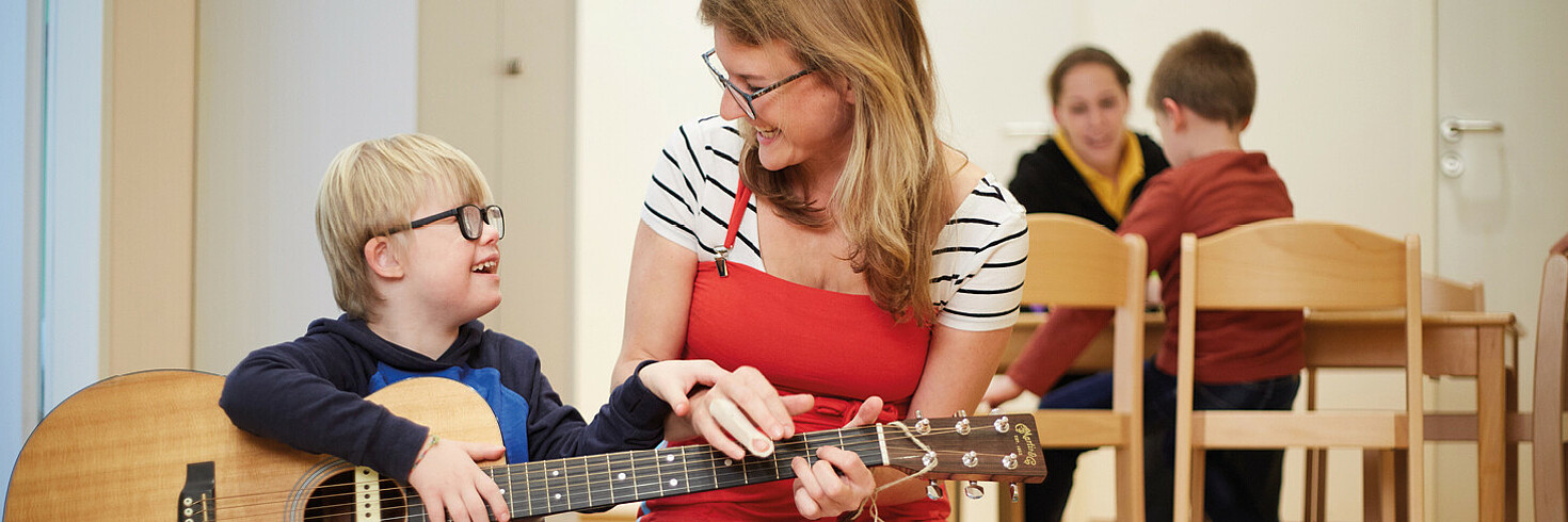 Ein blondes Kind hält eine Gitarre und lächelt eine Frau an, die dem Kind lächelnd dabei hilft, die Gitarre zu spielen